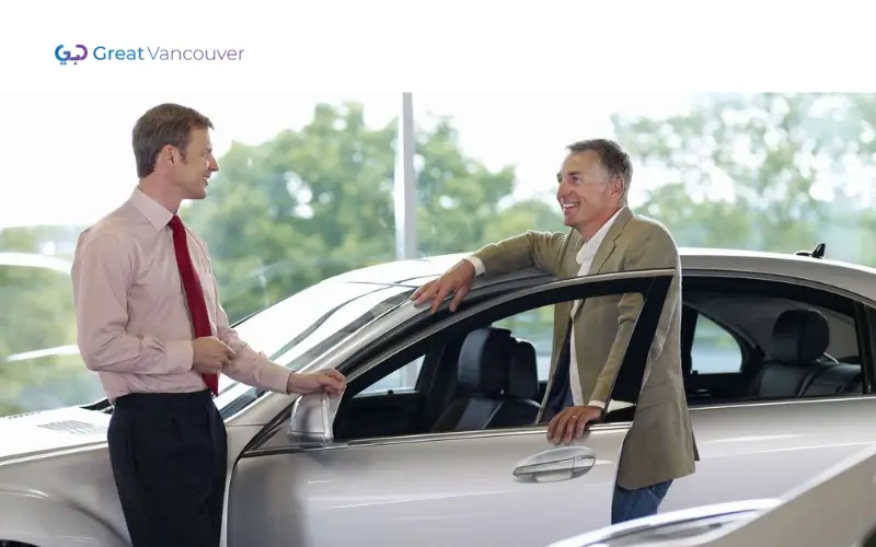 Two men are having a discussion next to a silver car in a showroom, with the 'Great Vancouver' logo in the background, as they explore exciting opportunities in Automobile Sales Jobs.